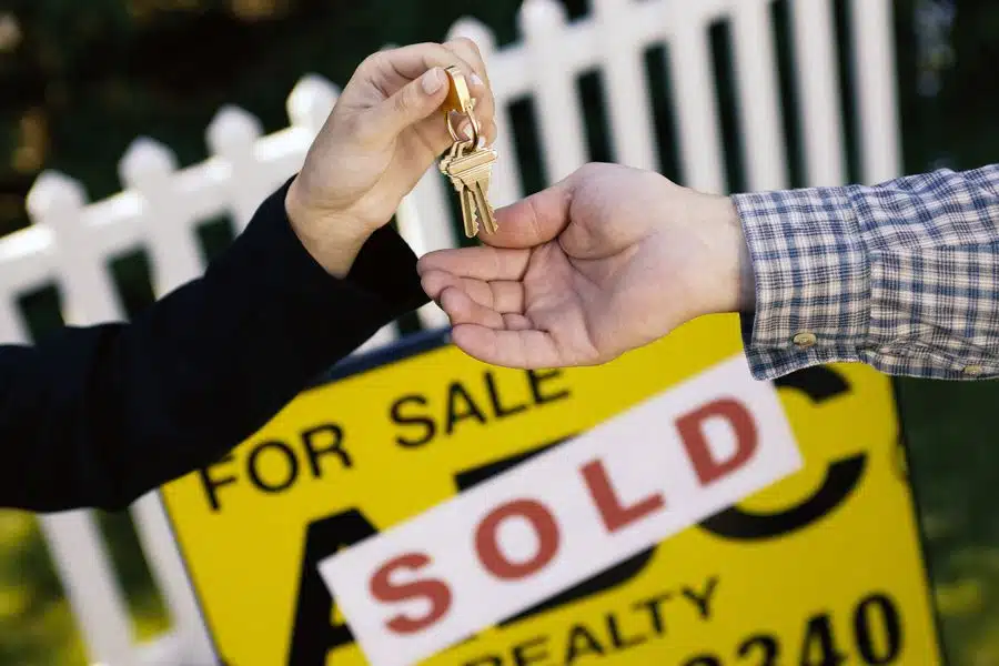 Woman handing man keys in front of house sold sign